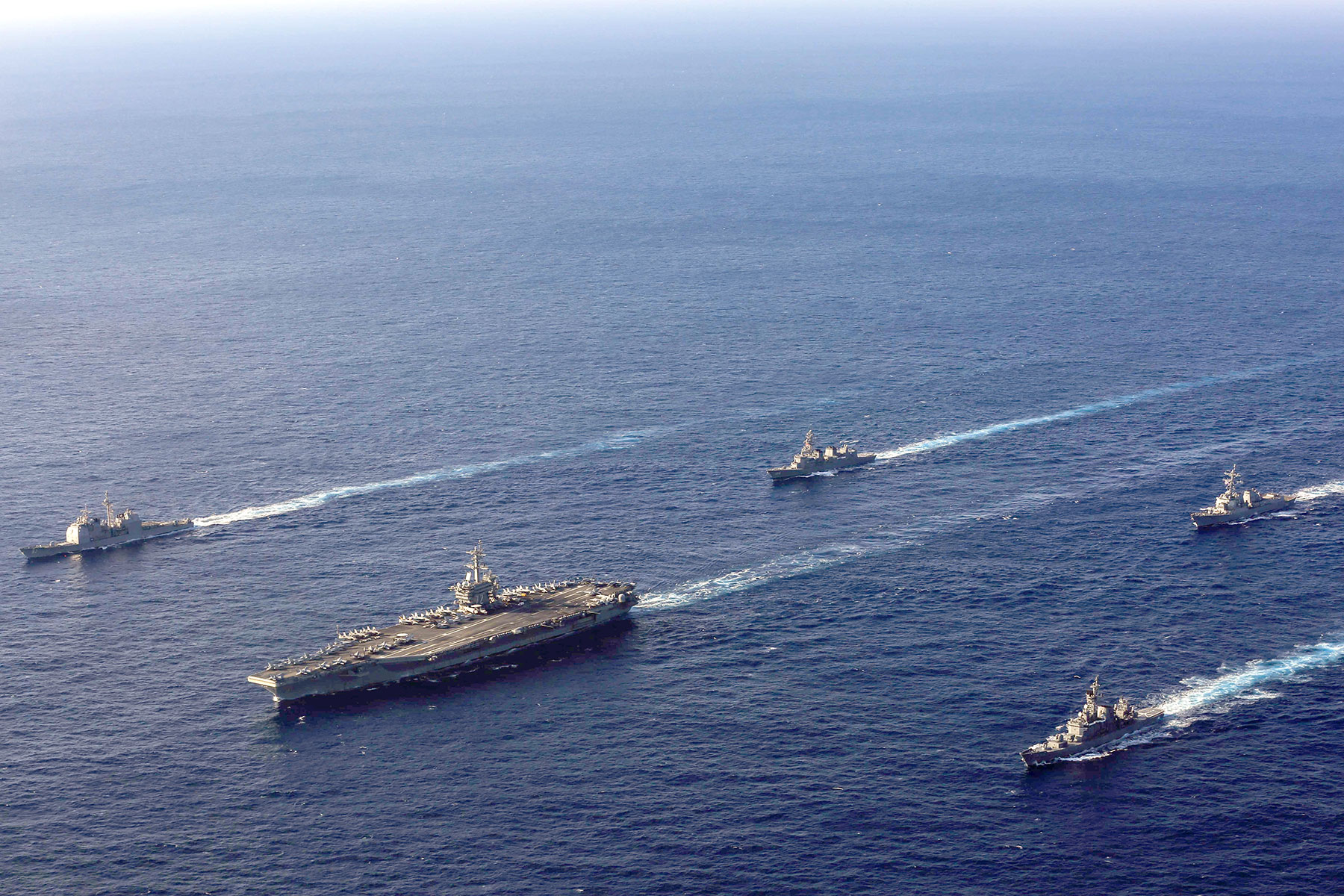 An image of the ocean and a fleet of Navy ships, one very large ship and four small ones moving in the water what appears at full speed.