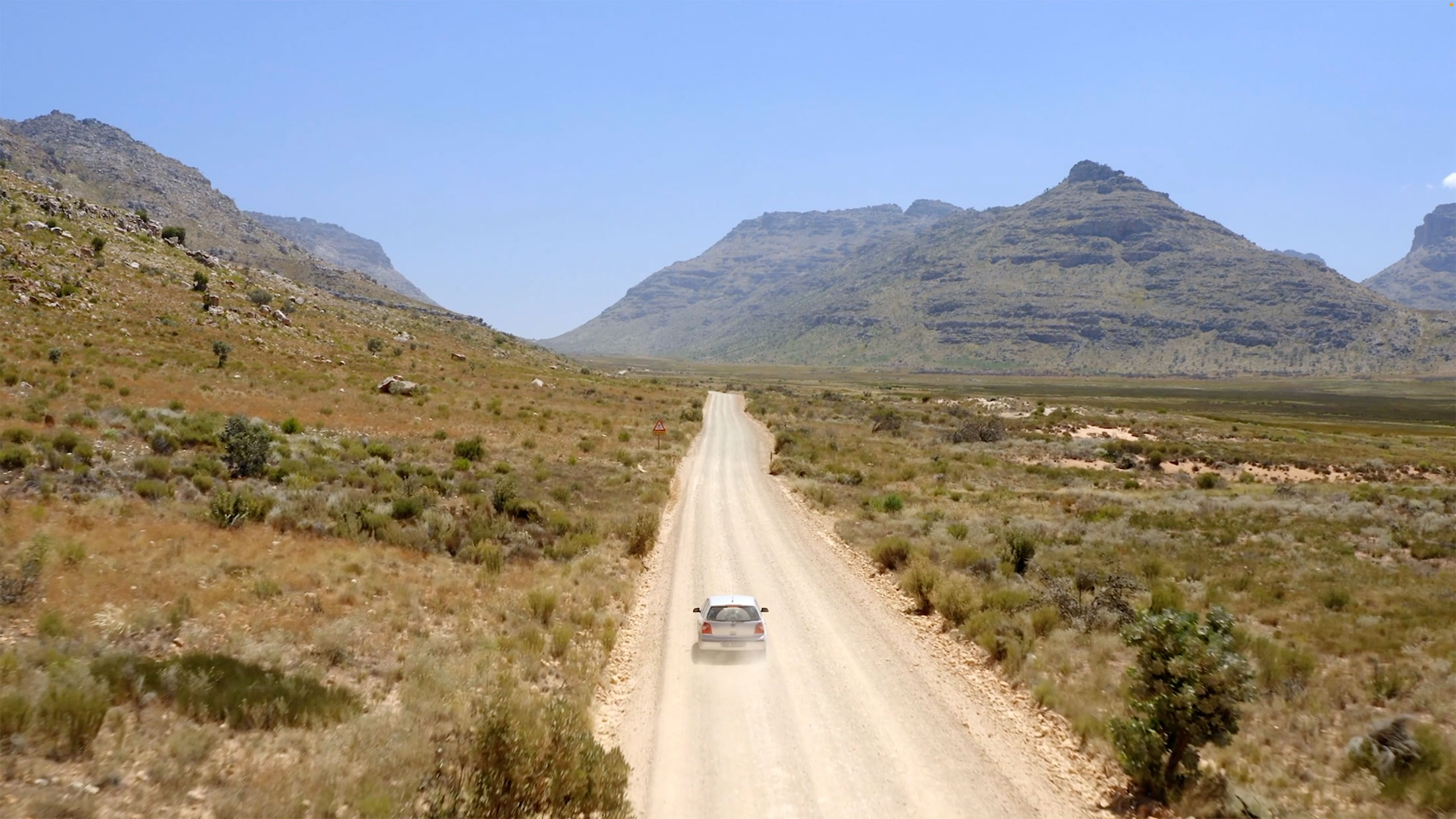 a silver car driving down a long straight dirt road in a remote desert landscape with large hills or mountains in the background.