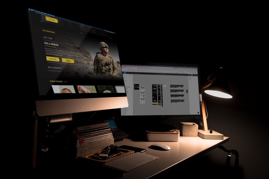 An image of a dark room with two computer monitors on a desk showing web design work and figma mockups for Army National Guard Legacy Campaign.
