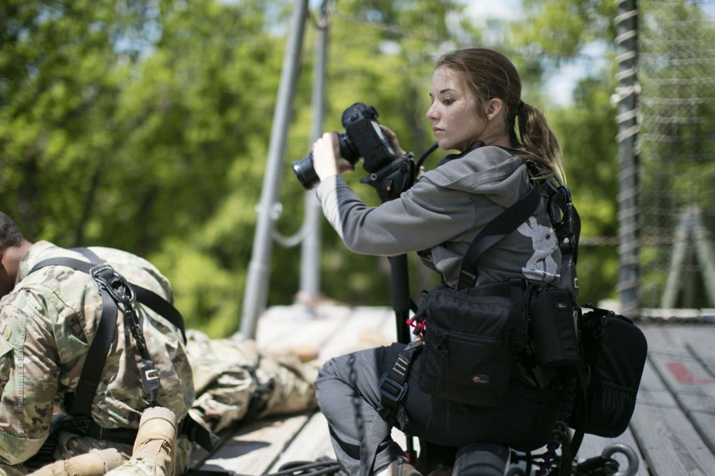 an image of a lady holding a camera and camera equipment behind the scenes of a video shoot with a national guard soldier in the background.