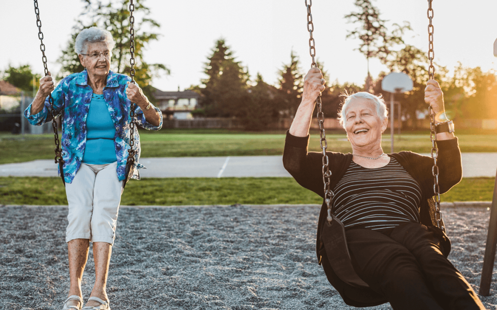 Two elderly ladies swinging on swings in a park.